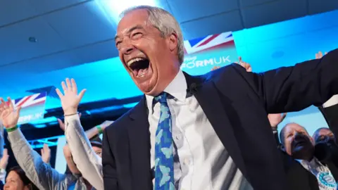 Nigel Farage - a man with silver hair and dressed in a suit with blue tie - laughs happily onstage while surrounded by supporters