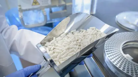 Getty Images White capsules in a silver shovel in a laboratory being held by a person in a white coat with blue gloves.