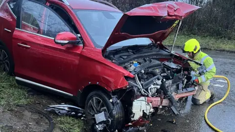Supplied A red car with severe front-end damage rests on a muddy roadside verge, while emergency personnel work at the scene beside a wet road.