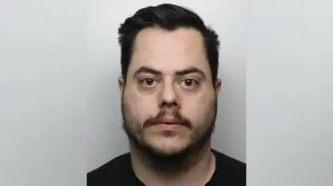 The image shows a close-up, front-facing portrait of a person against a plain light-grey background. The person has short dark hair, facial hair around the jaw and upper lip, and is wearing a dark-coloured top. The photograph is evenly lit and resembles an identification or mugshot-style image with the subject centred in the frame.