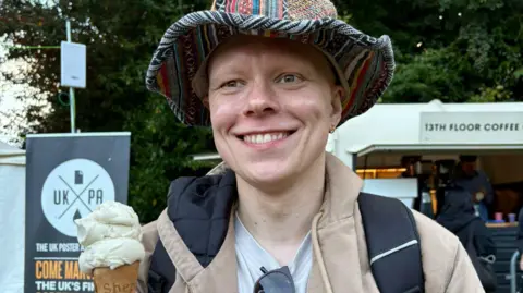 A man is smiling and holding a vanilla ice cream in his right hand, looking straight ahead. He is wearing a beige raincoat and a multi-coloured fabric hat with a wide brim.