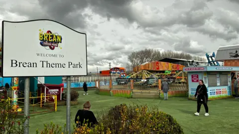 A sign reads 'Welcome to Brean Theme Park' with the park's logo on it. Some fairground rides are visisble in the background. People walk about on astroturf. It is a grey, cloudy day.