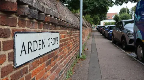 Christopher Day/LDRS A brick wall with a street sign saying "Arden Grove" next to pavement and a row of parked cars