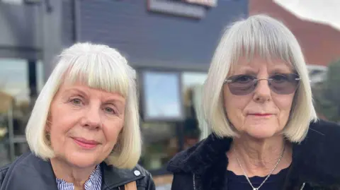 Penny Brind and Nicky Bettridge both have light-grey hair in a bob style with a fringe. Ms Brind wears a leather jacket over a striped top and Ms Bettridge, who is taller, has dark glasses and a dark blue jacket or coat.