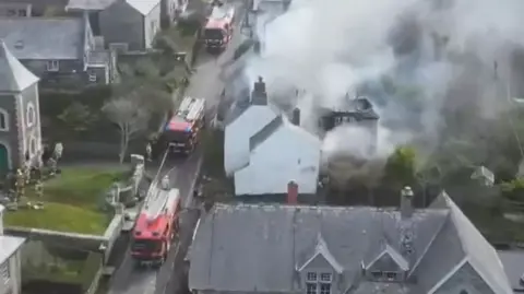 Fire trucks on a road next to a building which is on fire. There is smoke billowing from the roof of the building. There are houses lining either side of the street.
