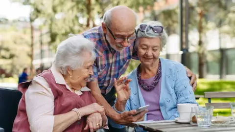 Getty Images Three older people beside each other looking at a phone the man is holding. Both women have grey hair and the man is bald with short grey hair at the side.