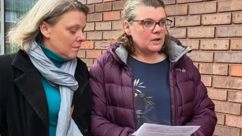 Two women standing next to each other. The woman on the left has blonde hair and is wearing a blue top, grey scarf and black jacket. The woman on the right has brown hair and is wearing glasses, a purple coat and a dark blue top.