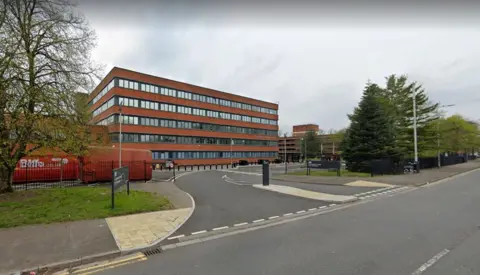 Google The outside of Companies House on Crown Way - the building is brown with multiple rows of windows and there are grassy patches and trees to either side and a pavement and road in front.