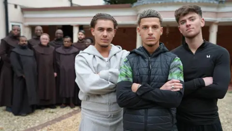 Three young men standing with their arms crossed with a group of traditional friars standing behind them in a courtyard.