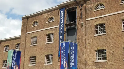 Exterior of the Museum in Docklands, a large brick warehouse building with barred windows and blue vertical banners hanging from the facade.