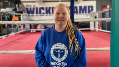 Simon Thake/BBC A woman in a blue sweatshirt smiles as she stands in front of a boxing ring with a red canvas.