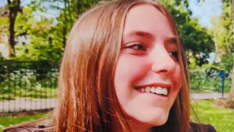 Family photo of Chloe Longster smiling in a park. You can see his head and shoulders. She wears her school uniform and has long light brown hair. She looked to her left.