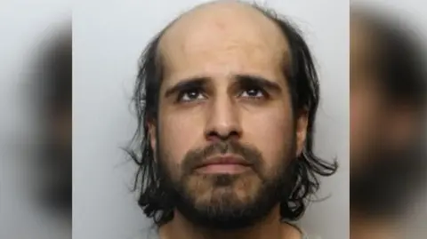 Police mugshot of a man with balding grey hair wearing a grey t-shirt