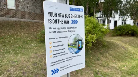 George Carden/BBC A sign by a bus stop which says "your new bus shelter is on the way" and has a picture of a new bus stop 
