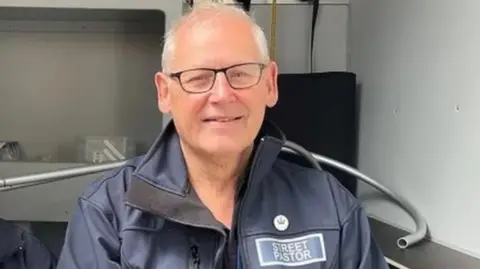 A man with white hair and glasses in a dark blue coat in a grey room with a silver metal tube behind him
