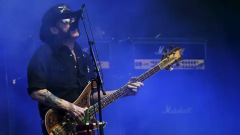 Reuters Lemmy Kilmister - wearing a black hat and black shirt - sings into a microphone while he plays a bass guitar on stage. The stage has been bathed in blue light and Marshall amplifiers can be seen behind him.