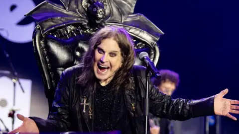 Ozzy Osbourne performing while seated on an elaborate black throne, which was topped by a giant black bat. His eyes and mouth are widened while is hands spread out to the side

