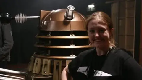 Jane Lutwyche Jayne with tied back brown hair wearing a black Doctor Who t-shirt, stood in front of a model Dalek
