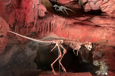 Two replica skeletons sit in alcoves in Kents Cavern lit by atmospheric red light.