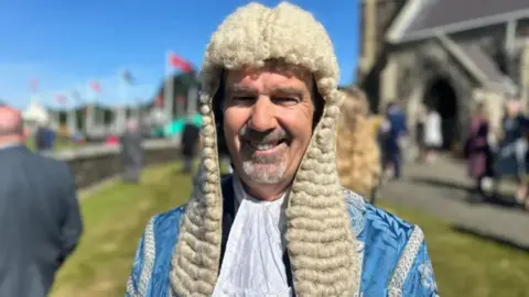 BBC Laurence Skelly is wearing light blue ceremonial robes and a traditional parliamentary wig. He has a grey goatee beard and is smiling while standing outside a chapel. Rows of Manx flags are flying on a sunny day.