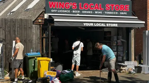 Ollie Bayliss/BBC Three people outside a shop, helping to clear it up. One is holding a broom, another person is by some bins. There are lots of items on the ground and by bins. The shop front has items in it, but it is dark. 