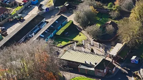 Roarr! An aerial image of long, rectangular agricultural buildings, fenced-off grass areas, groups of trees and paved areas. 
