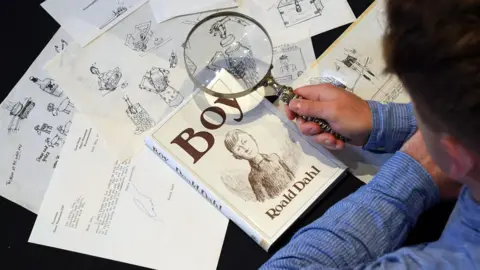 An aerial image showing sketches of illustrations and a copy of 'Boy' written by Roald Dahl. A person is holding a magnifying glass up to the copy of Boy. The person is wearing a blue shirt.