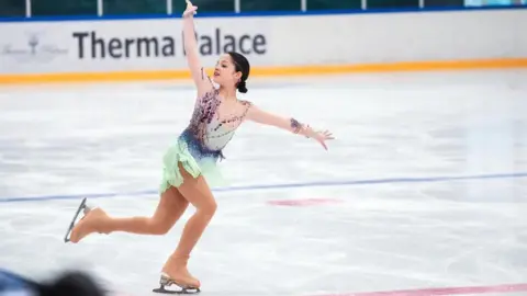 BRITISH ICE SKATING Leilah is figure skating on the ice with her arms outstretched and one leg risen behind her, smiling.