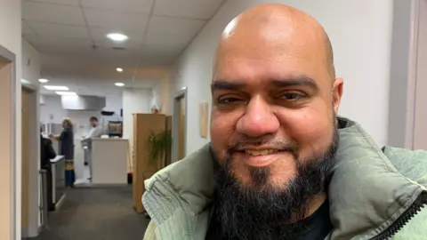 Arif Voraji, wearing a green coat, smiling at the camera. He has a dark beard.