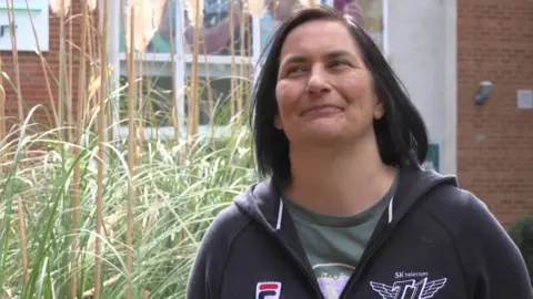 Jane who has shoulder length straight dark brown hair and is smiling, is wearing a black Fila hooded zip up sweatshirt with a green T-shirt underneath. She is standing in front of a red bricked building and a large pampas grass bush./ 
