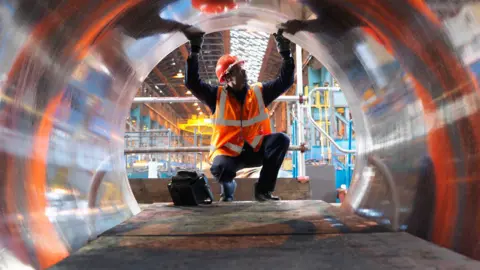Engineer wearing an orange high-vis jacket and orange helmet testing forged steel in a factory in Sheffield, UK