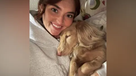Jess McKee wearing a grey tracksuit cuddles a sandy-coloured dog. She is smiling. 