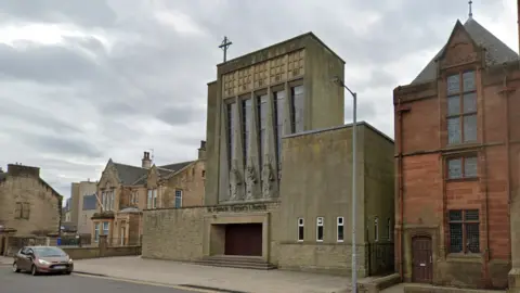 Google St Francis Xavier Catholic Church in Falkirk. The building is dark stone and has a cross on top of it. A red brick building is to the right and a brown Ford car is parked in front.