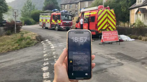 A phone screen with an emergency alert on is seen on a phone, with fire engines behind