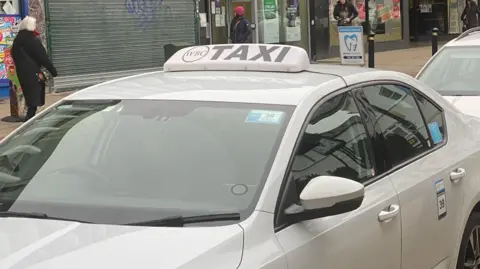 Two white taxis are shown parked in a straight line, with shops on a high street in the background, with people walking in both directions.