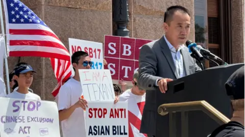 Ning Li Jason Yuan gives a speech at a lectern, with protesters behind him holding up signs