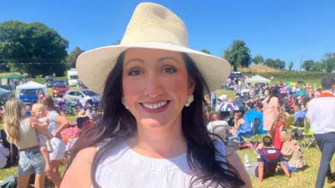 A woman with long brown hair is smiling and looking at the camera. She is wearing a light coloured straw hat and a white sleeveless top. There are crowds of people in the background standing and sitting in a field.