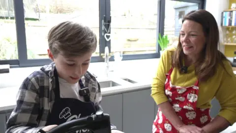 Austin and BBC presenter Jo Kent stand behind a kitchen island wearing aprons,. Austin is working with a mixer, while Jo stands next to him and smiles. Austin has wavy brown hair and wearing a black and white checked shirt. Joe wears a yellow jumper with a red apron.