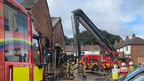 Firefighters inside a cordon tackling a fire at a storage unit on an industrial estate with two fire engines one with an aerial platform close by and a row of houses nearby.