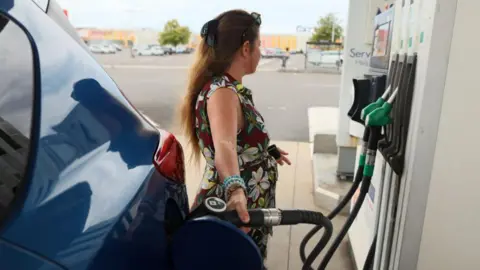 A woman in a floral dress fills her car at a petrol station.
