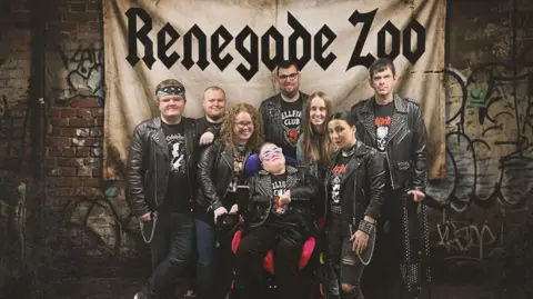 Marty Doherty The members of Derry band renegade Zoo are dressed in black leather jackets. There are eight people, four men and four women. In centre of the pic is a woman in a wheelchair. The others are stood around her. In the background is a banner with the band's name written on it