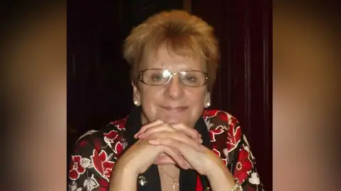 Hayley Charman A woman with glasses, a red top and her head leaning on her clasped hands. She is smiling softly at the camera.