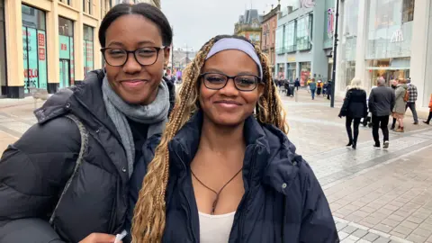 Alex Moss/BBC Two smiling women stood in a city centre precinct. Both are wearing glasses. The woman on the left has black hair pulled back and is wearing a black padded jacket and grey scarf. The other woman has a white vest with a navy padded jacket in. She has long light brow hair with braids and is wearing a lilac headband. 