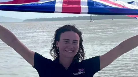 Zara is holding a large Union Jack flag high above her head. She is smiling at the camera and wears a blue Team Forces branded polo shirt. Behind her is the sea. Zara has brown curly hair that looks slightly wet at the ends from the rowing. 