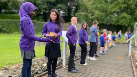 Schoolchildren in purple uniform are standing in a line on a walkway with grass and trees behind them. Two girls closest on the left are handing the baton over to the other.