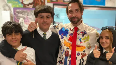 Three teenagers smile towards the camera, they are wearing white shirts and green ties and are putting their thumbs up. A man with floppy brown hair and a beard stands between them, he is wearing a white shirt with colourful graphics on.