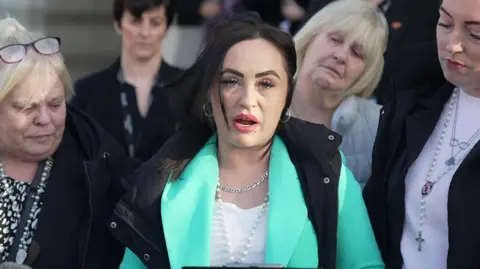 Antonia Gawith looks at the camera a she reads a statement outside court. She is wearing a white top and black sleeveless coat. She has dark brown hair and silver hoop earrings. She is surrounded by a four other women.