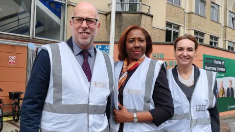Three people are standing in front of Sir Charles Frossard House. All three are wearing white vests over their formal clothes. On the left is a white man with a bald head, glasses and a grey beard. In the middle is a black woman with auburn hair. On the right is a white woman with brown hair. 