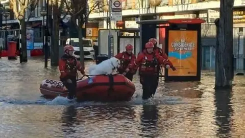 A street lines with houses and flooded. A boat is being led down the road by three men in high viz. A dog is in the boat.