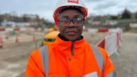 Happy Konete wearing an orange hi-vis jacket, glasses and hard hat with The Skills Centre written on it. A blurred building site and red and white metal fencing is behind her